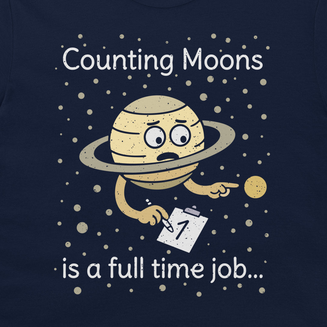 A print on a dark navy blue t-shirt featuring a cartoon of a sad, tired Saturn-like planet holding a clipboard with the number 1, surrounded by white text that reads 'Counting Moons is a full time job.