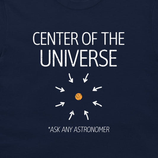 A close-up of a navy blue t-shirt featuring a white graphic print that reads 'CENTER OF THE UNIVERSE' with arrows pointing toward a small orange planet, and the text '*ASK ANY ASTRONOMER' below.
