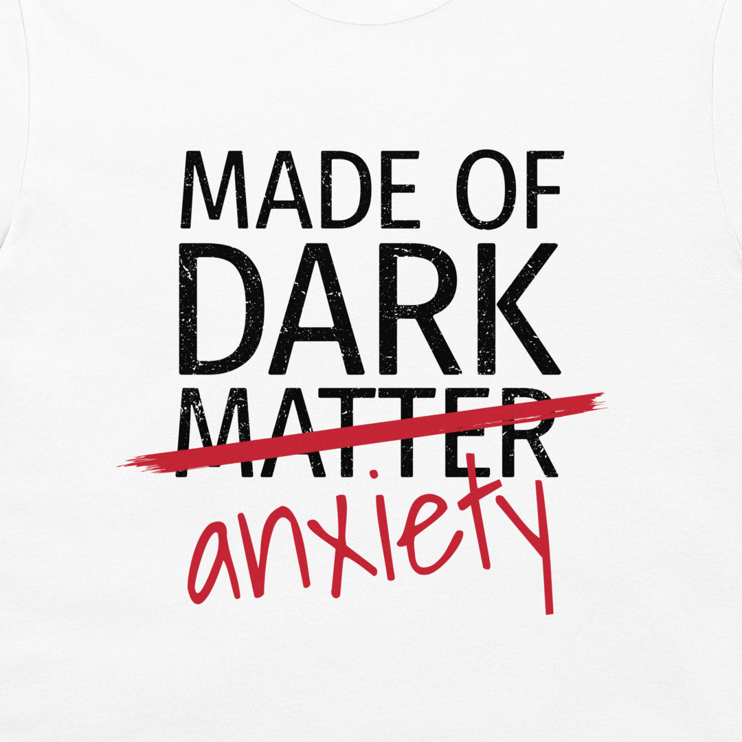 White t-shirt featuring a graphic print with the text 'MADE OF DARK MATTER' in distressed black block letters, crossed out with a red brush stroke, and 'anxiety' written below in red script.