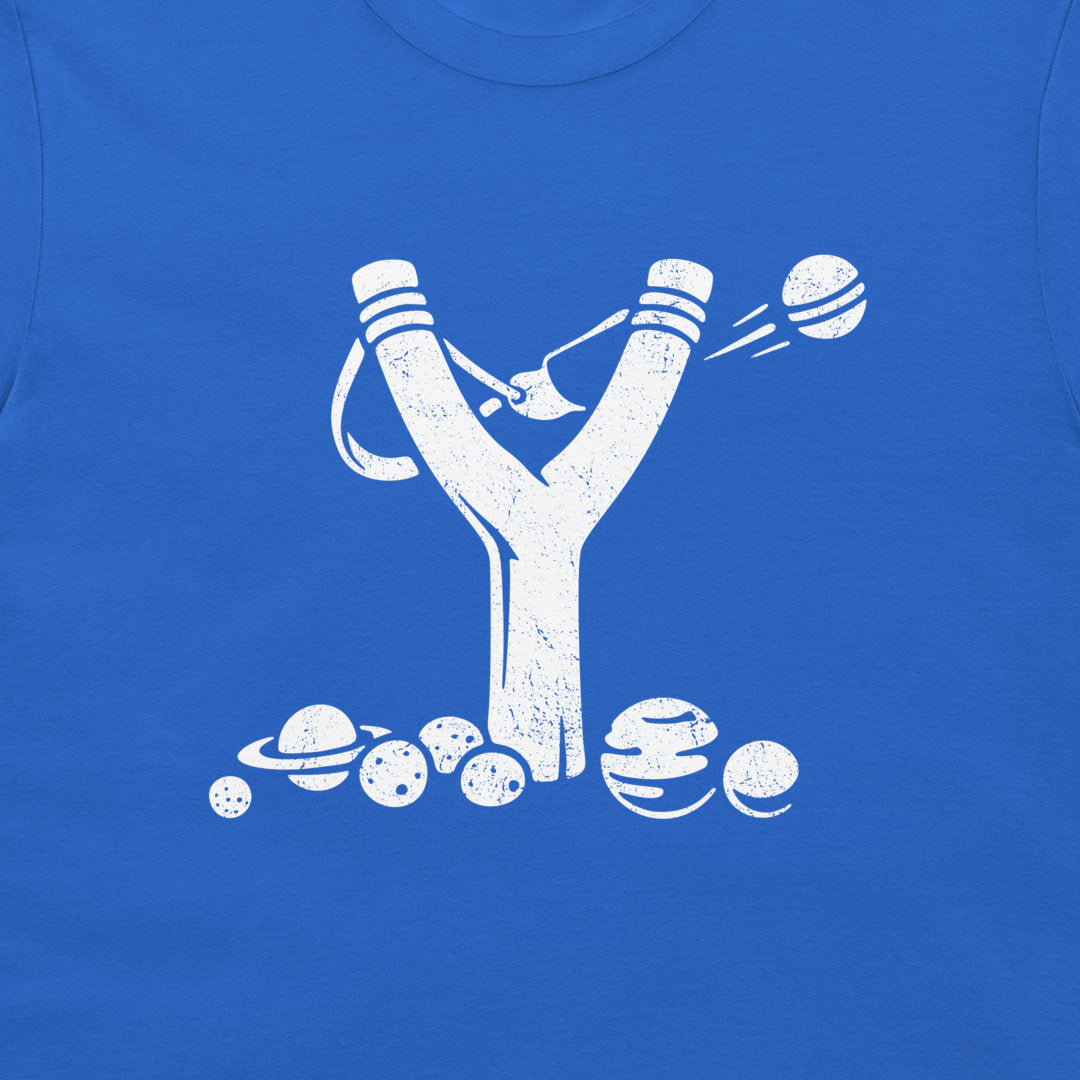 A close-up of a royal blue T-shirt featuring a distressed white graphic of a slingshot launching planets and moons, with several planets and moons illustrated along the bottom.