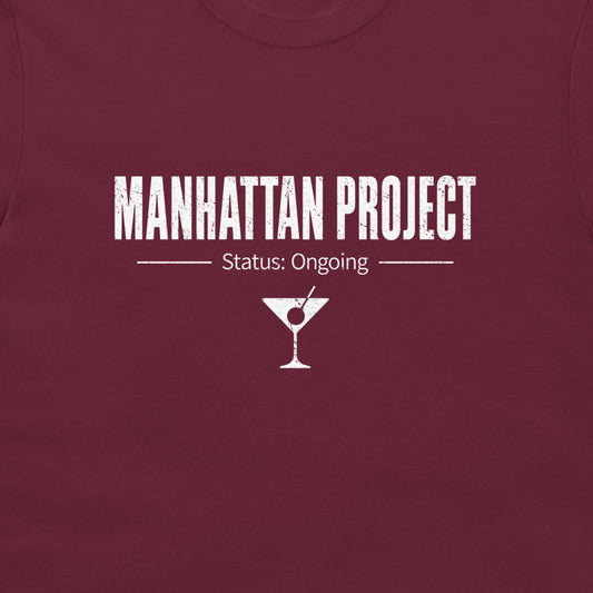 A close-up of a maroon crewneck t-shirt featuring a white distressed graphic that reads 'MANHATTAN PROJECT' with 'Status: Ongoing' and a martini glass icon below.