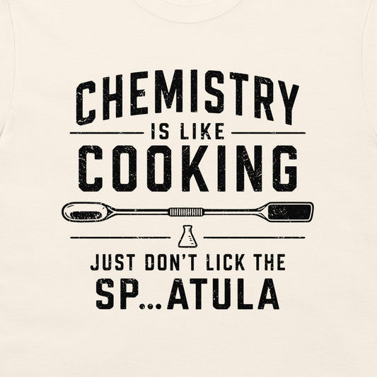 A close-up of a light cream-colored t-shirt featuring a black distressed graphic print that reads 'CHEMISTRY IS LIKE COOKING' with a spatula illustration and 'JUST DON'T LICK THE SP...ATULA'.