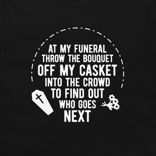 A close-up of a black t-shirt, featuring a white graphic print with the text 'AT MY FUNERAL THROW THE BOUQUET OFF MY CASKET INTO THE CROWD TO FIND OUT WHO GOES NEXT' along with a coffin and flowers icon.