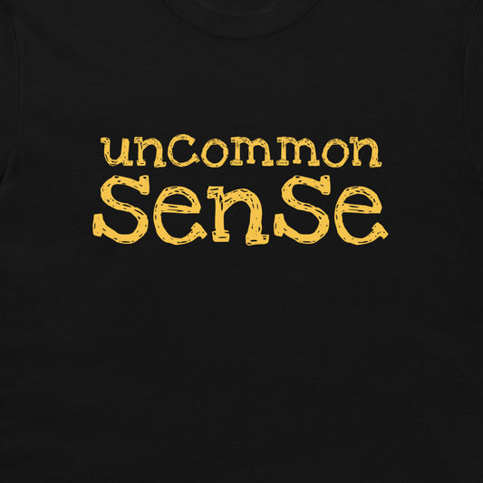 A close-up of a black t-shirt featuring the phrase 'uncommon sense' printed across the chest in a textured, distressed yellow font.
