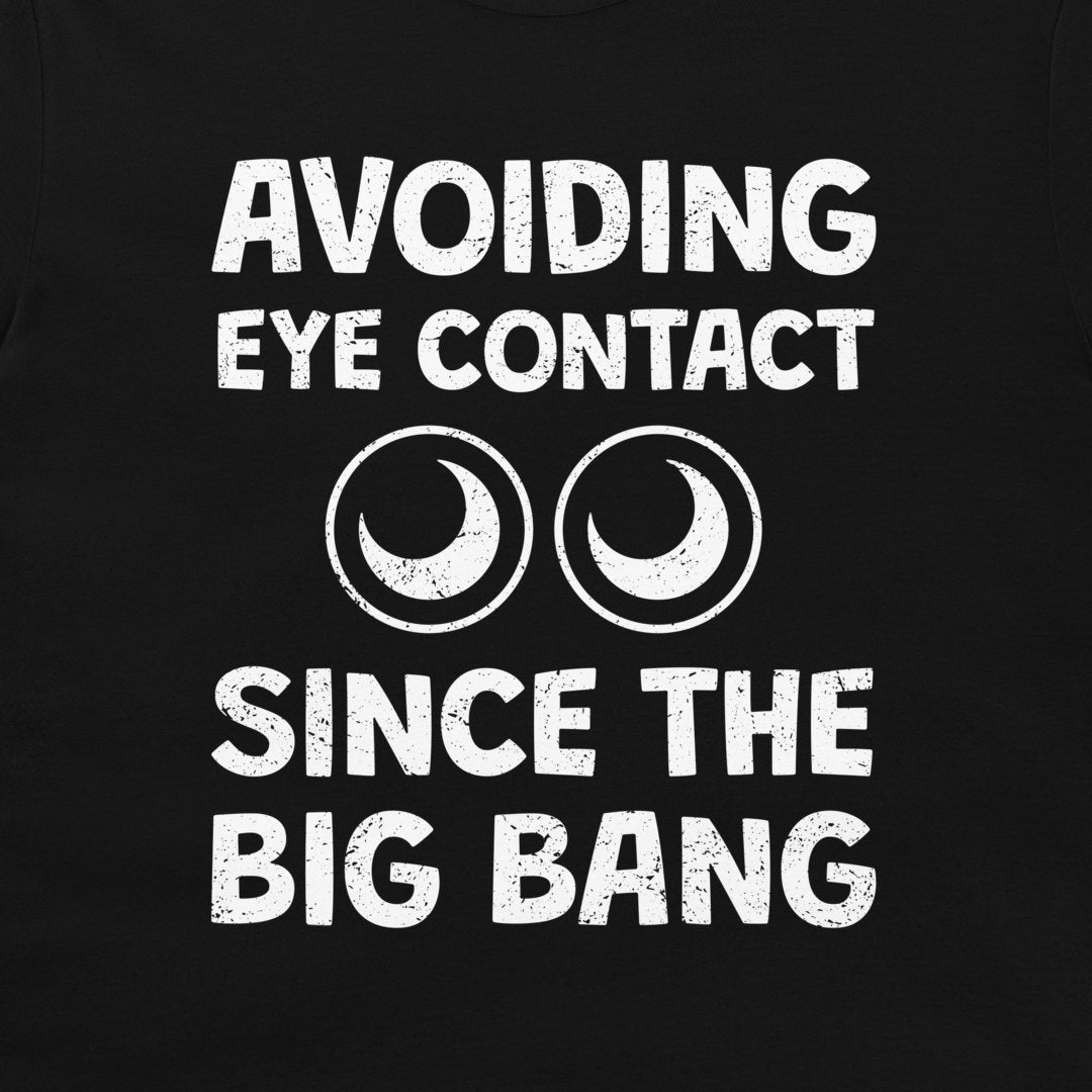 A close-up of a black garment, likely a t-shirt, featuring a distressed white graphic print that reads 'AVOIDING EYE CONTACT SINCE THE BIG BANG' with two crescent moon and circle eye symbols.