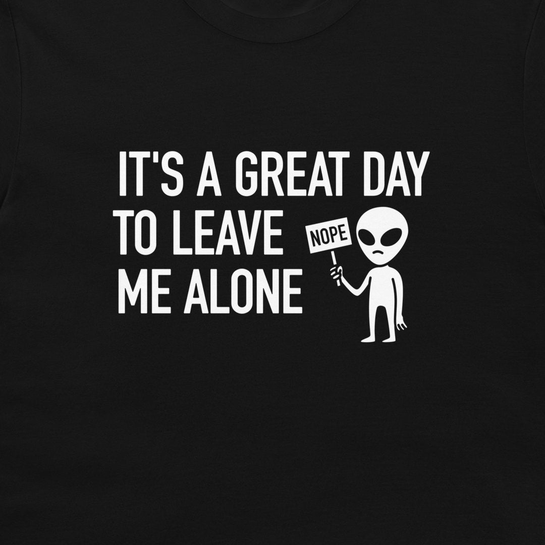 A close-up of a black t-shirt featuring a white graphic print that reads 'IT'S A GREAT DAY TO LEAVE ME ALONE' next to a cartoon alien holding a 'NOPE' sign.