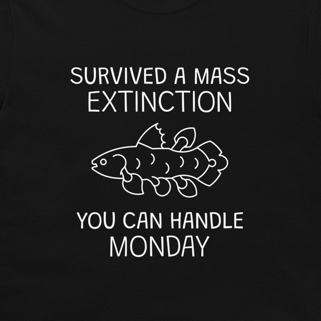 A close-up of a black garment, likely a t-shirt, featuring a white graphic print. The design displays the text 'SURVIVED A MASS EXTINCTION YOU CAN HANDLE MONDAY' surrounding a simple white line drawing of a fish.