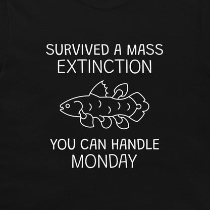 A close-up of a black garment, likely a t-shirt, featuring a white graphic print. The design displays the text 'SURVIVED A MASS EXTINCTION YOU CAN HANDLE MONDAY' surrounding a simple white line drawing of a fish.