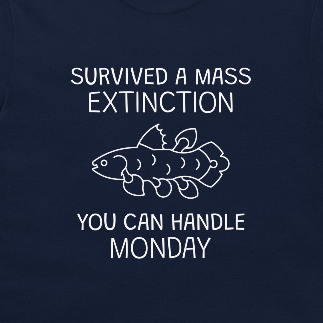 A close-up of a navy blue garment, likely a t-shirt, featuring a white graphic print. The design displays the text 'SURVIVED A MASS EXTINCTION' and 'YOU CAN HANDLE MONDAY' surrounding a simple white line drawing of a fish.