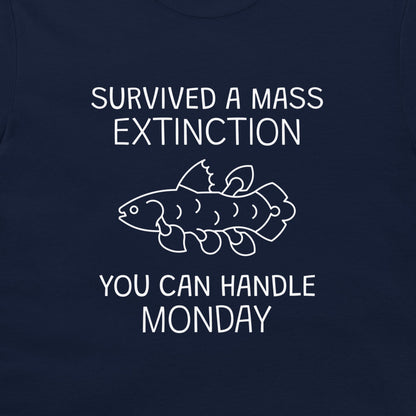 A close-up of a navy blue garment, likely a t-shirt, featuring a white graphic print. The design displays the text 'SURVIVED A MASS EXTINCTION' and 'YOU CAN HANDLE MONDAY' surrounding a simple white line drawing of a fish.