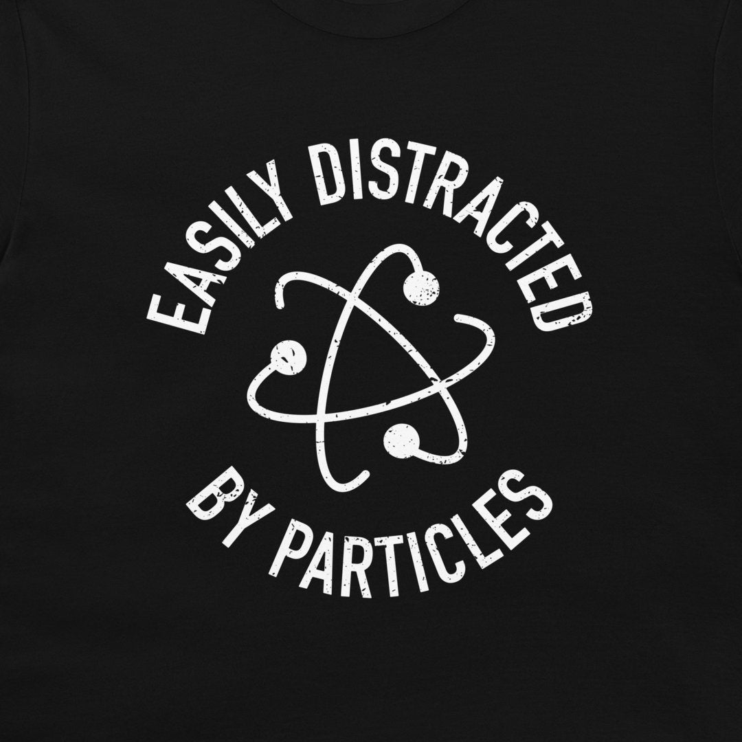 A close-up of a t-shirt, featuring a distressed white graphic print of an atom symbol with the text 'EASILY DISTRACTED BY PARTICLES'.