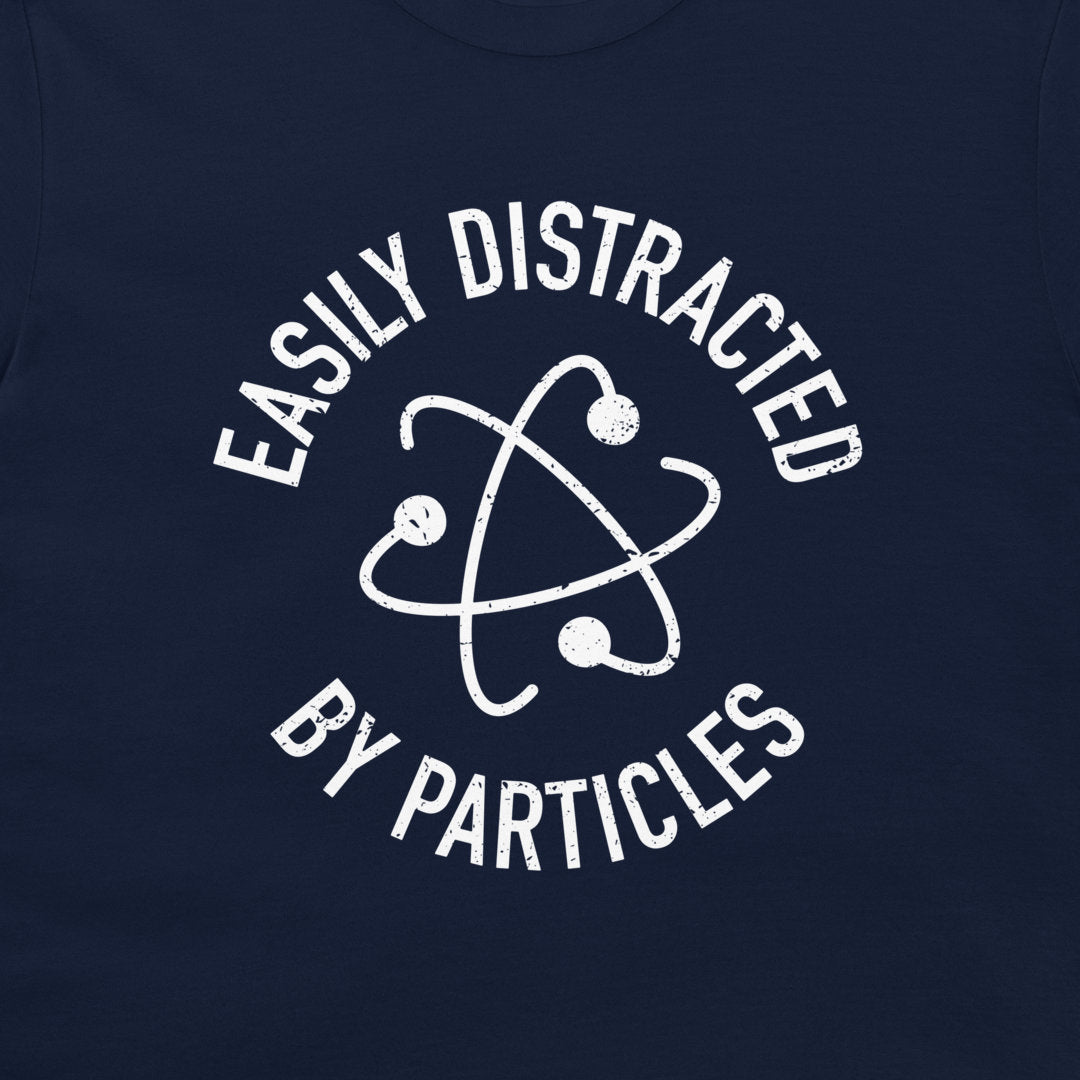 A close-up of a navy blue t-shirt, featuring a distressed white graphic of an atom symbol with the text 'EASILY DISTRACTED BY PARTICLES'.