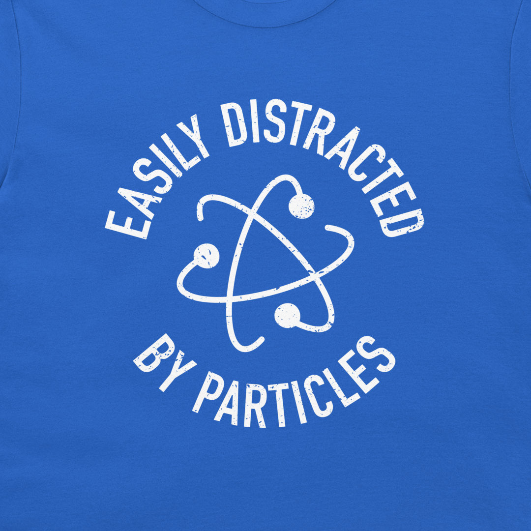 A close-up of a royal blue t-shirt featuring a white distressed graphic of an atom symbol with the text 'EASILY DISTRACTED BY PARTICLES'.