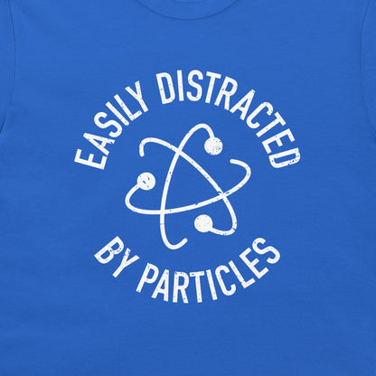 A close-up of a royal blue t-shirt featuring a white distressed graphic of an atom symbol with the text 'EASILY DISTRACTED BY PARTICLES'.