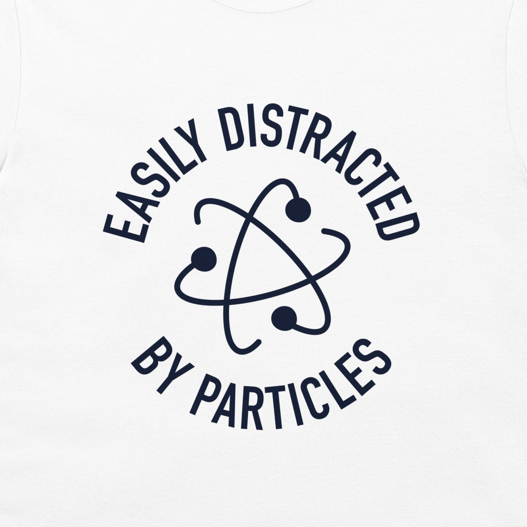 A close-up of a white t-shirt, featuring a dark navy blue graphic print. The design displays the text 'EASILY DISTRACTED BY PARTICLES' in a circular layout surrounding an atom symbol.
