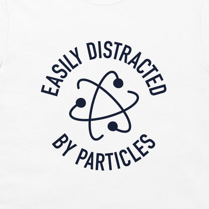 A close-up of a white t-shirt, featuring a dark navy blue graphic print. The design displays the text 'EASILY DISTRACTED BY PARTICLES' in a circular layout surrounding an atom symbol.