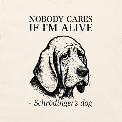 A close-up of a cream-colored t-shirt featuring a black graphic print of a sad-looking bloodhound dog with the text 'NOBODY CARES IF I'M ALIVE' and '- Schrödinger's dog'.