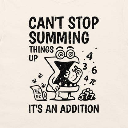 A close-up of a cream-colored t-shirt featuring a black graphic print. The design shows a cartoon letter 'X' character in a toilet bowl with the text 'CAN'T STOP SUMMING THINGS UP' above and 'IT'S AN ADDITION' below, surrounded by numbers and a calculator.