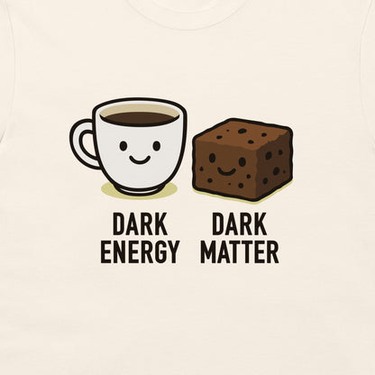 A close-up of a light cream-colored garment, likely a t-shirt, featuring a cartoon graphic of a smiling coffee cup labeled 'DARK ENERGY' next to a smiling brownie labeled 'DARK MATTER'.