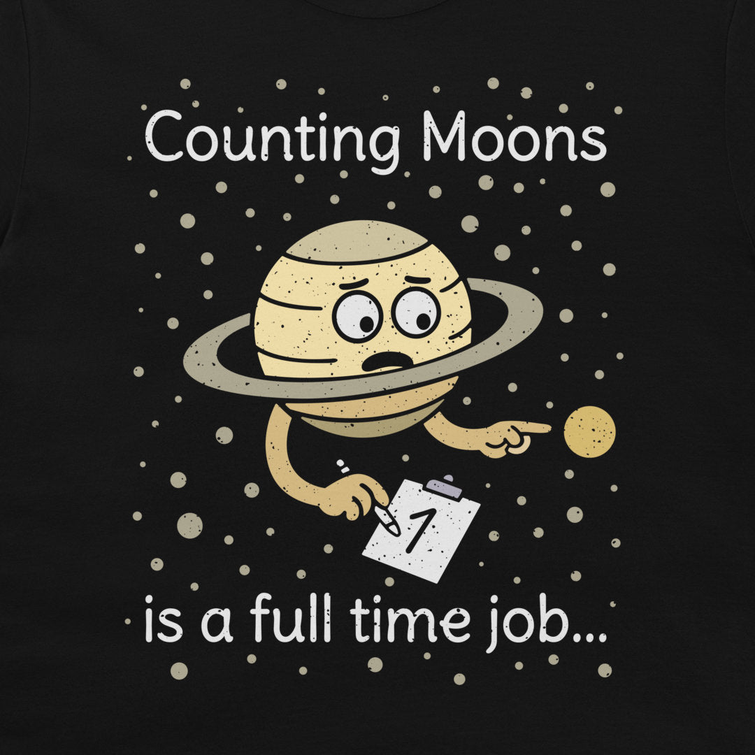 A print on a black t-shirt featuring a cartoon of a sad, tired Saturn-like planet holding a clipboard with the number 1, surrounded by white text that reads 'Counting Moons is a full time job.