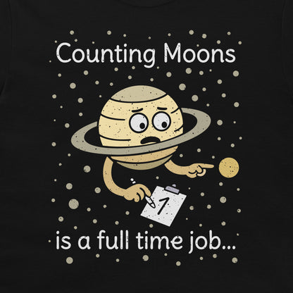 A print on a black t-shirt featuring a cartoon of a sad, tired Saturn-like planet holding a clipboard with the number 1, surrounded by white text that reads 'Counting Moons is a full time job.