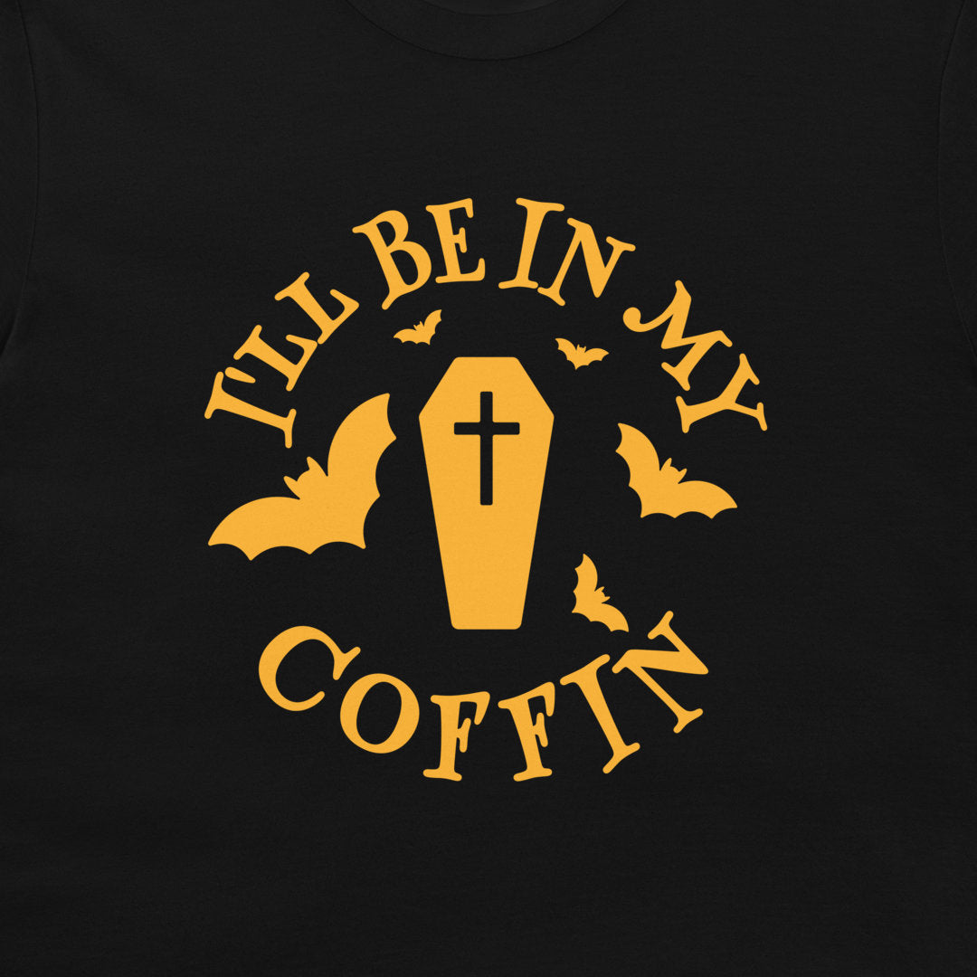 A close-up of a black t-shirt, featuring a yellow graphic print that reads 'I'LL BE IN MY COFFIN' with a coffin, bats, and a cross.
