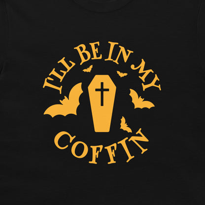 A close-up of a black t-shirt, featuring a yellow graphic print that reads 'I'LL BE IN MY COFFIN' with a coffin, bats, and a cross.