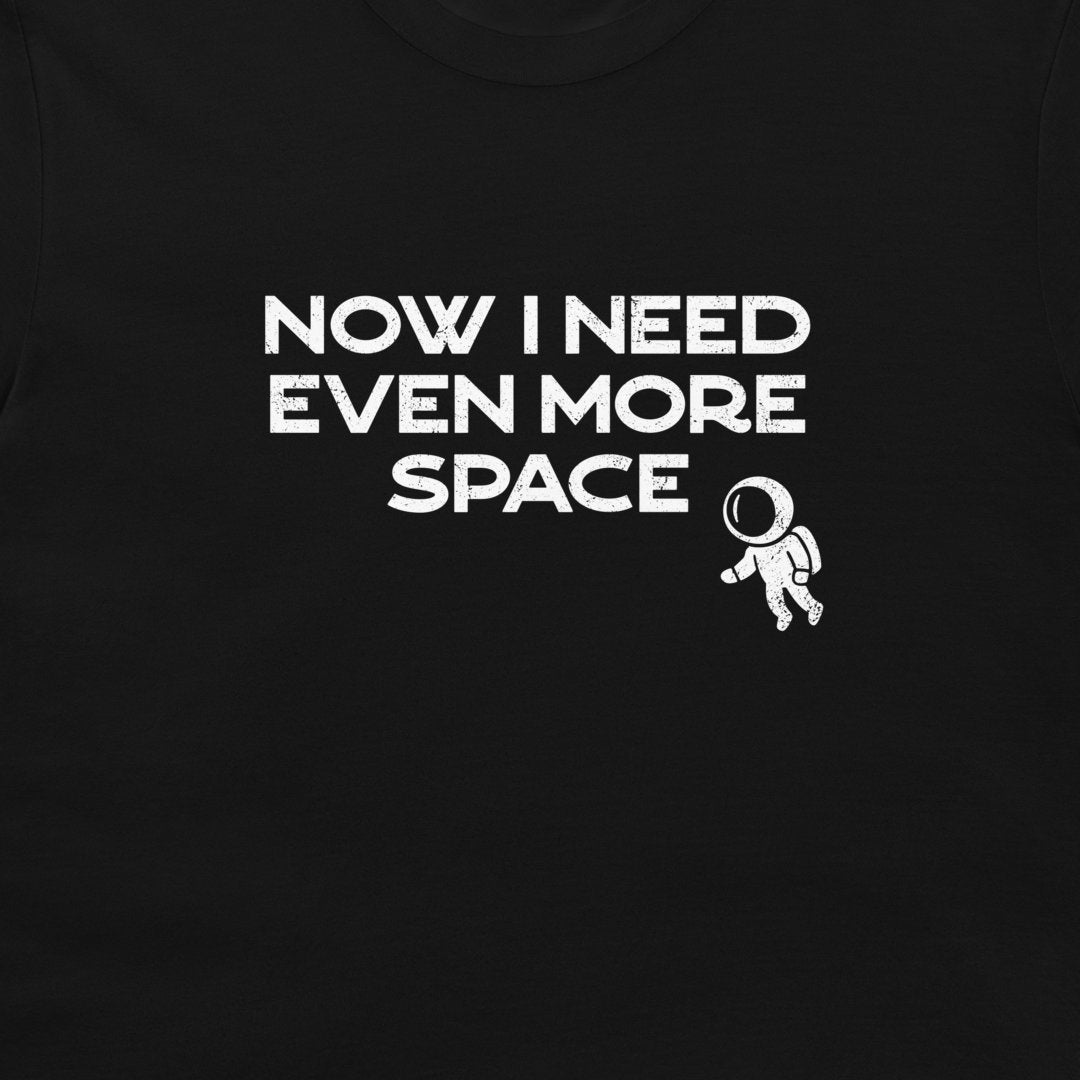 A close-up of a black t-shirt featuring a white graphic print that reads 'NOW I NEED EVEN MORE SPACE' next to a small astronaut illustration.