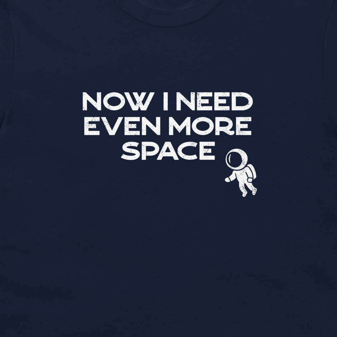 A close-up of a navy blue t-shirt featuring a white graphic print that reads 'NOW I NEED EVEN MORE SPACE' next to a small astronaut illustration.