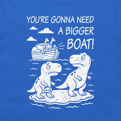 A close-up of a royal blue t-shirt featuring a white graphic print. The design shows two cartoon dinosaurs standing on a small island, with Noah's Ark depicted in the background under clouds and the text 'YOU'RE GONNA NEED A BIGGER BOAT!'.