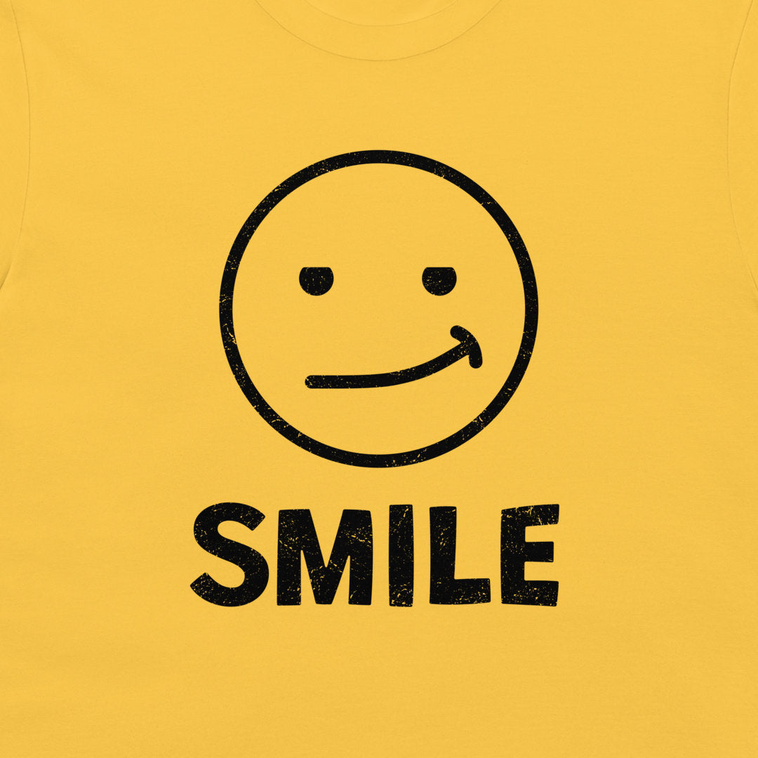 A close-up of a yellow t-shirt, featuring a black distressed graphic of a simple smiley face with the word 'SMILE' printed below it.
