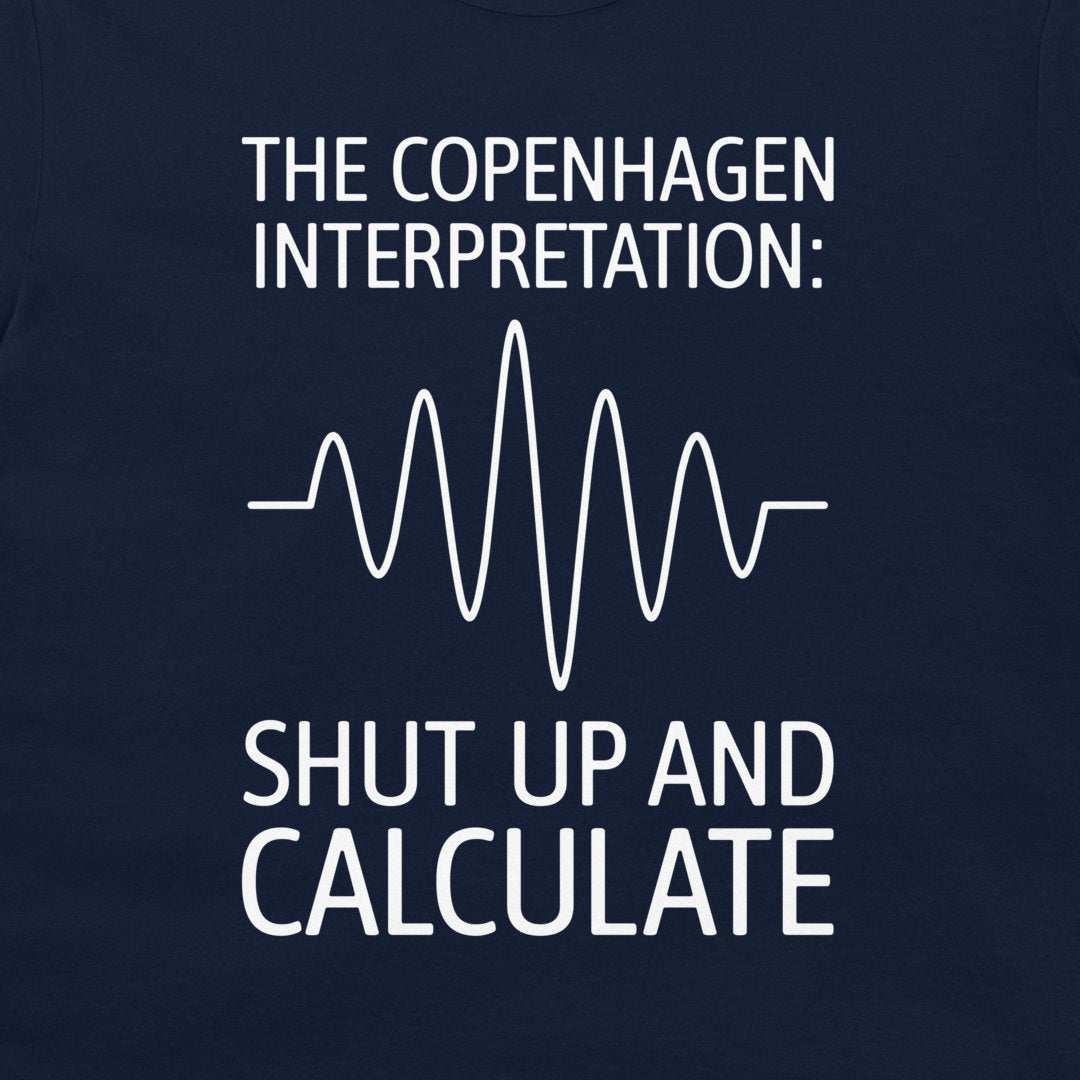 A close-up of a navy blue t-shirt featuring a white graphic print with the text 'THE COPENHAGEN INTERPRETATION:' and 'SHUT UP AND CALCULATE' surrounding a white heartbeat-style waveform.