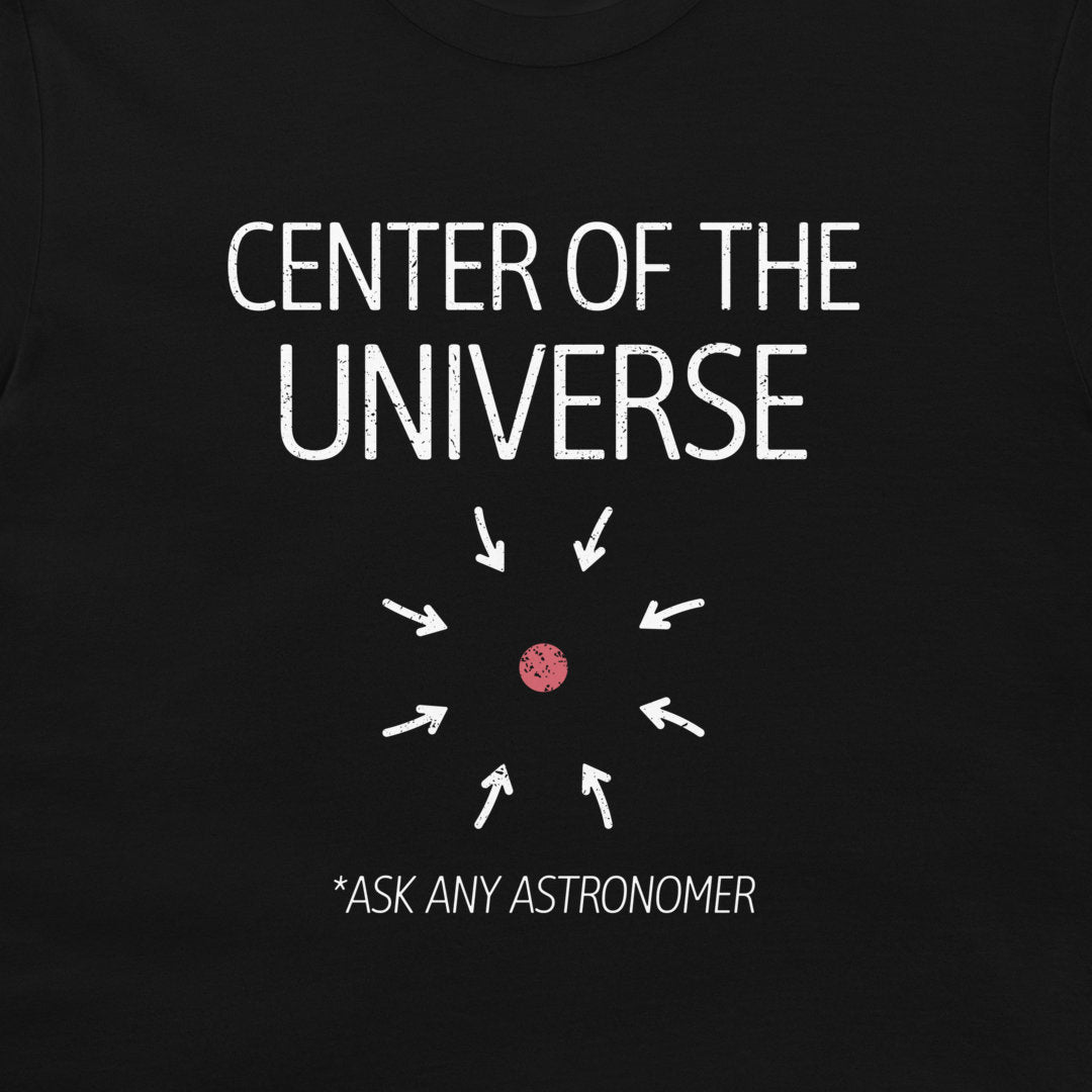A close-up of a black t-shirt featuring a white graphic print that reads 'CENTER OF THE UNIVERSE' with arrows pointing toward a small pink dot, and the text '*ASK ANY ASTRONOMER' below.