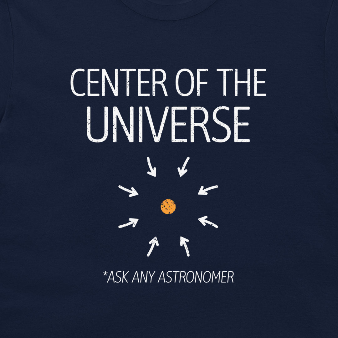 A close-up of a navy blue t-shirt featuring a white graphic print that reads 'CENTER OF THE UNIVERSE' with arrows pointing toward a small orange planet, and the text '*ASK ANY ASTRONOMER' below.