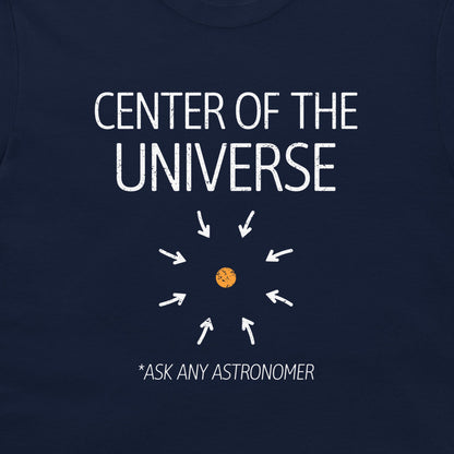A close-up of a navy blue t-shirt featuring a white graphic print that reads 'CENTER OF THE UNIVERSE' with arrows pointing toward a small orange planet, and the text '*ASK ANY ASTRONOMER' below.