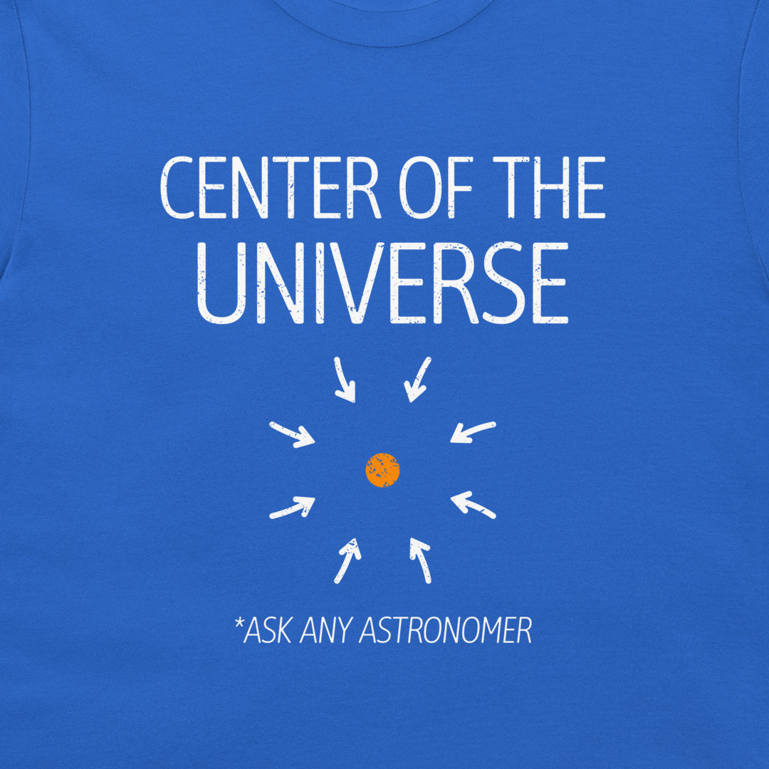 A close-up of a blue T-shirt with a white graphic print that reads 'CENTER OF THE UNIVERSE' above a small orange circle and white arrows, with '*ASK ANY ASTRONOMER' printed below.
