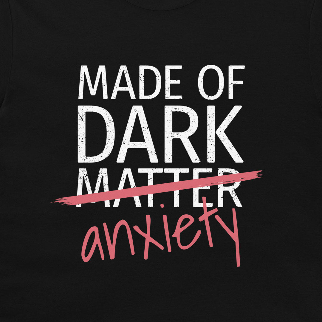 A close-up of a black graphic print on a shirt or garment. The design reads 'MADE OF DARK MATTER' in distressed white block letters, crossed out with a pink brushstroke, with 'anxiety' written below in pink script.