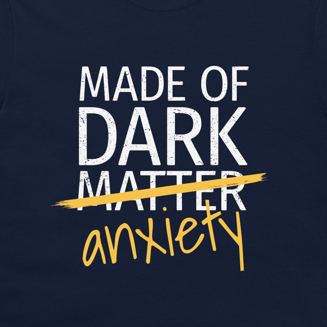 A close-up of a navy blue garment, likely a t-shirt, featuring a graphic print that reads 'MADE OF DARK MATTER' in distressed white block letters, crossed out with a yellow brushstroke, with 'anxiety' written below in yellow script.