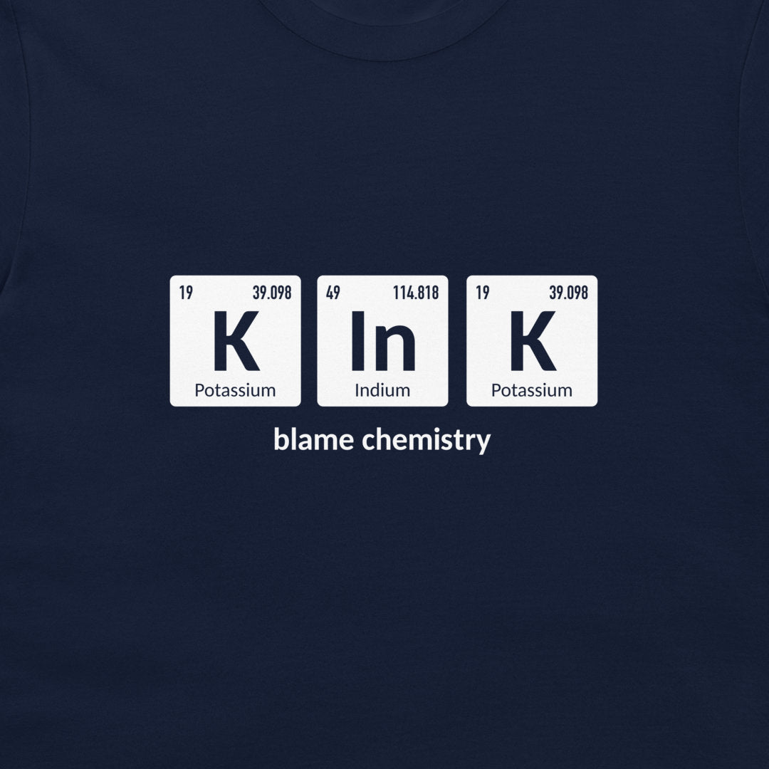 A close-up of a navy blue t-shirt featuring a white graphic print of three periodic table tiles spelling 'K In K' with the text 'blame chemistry' underneath.