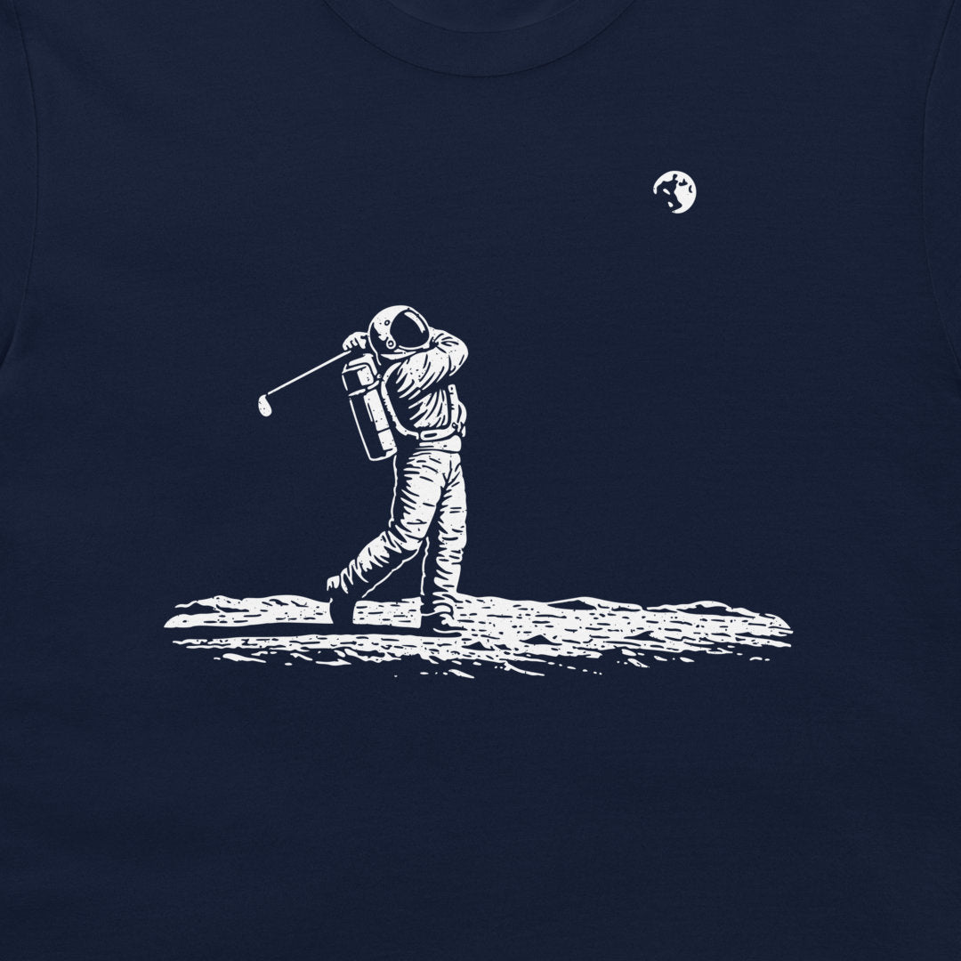A dark navy blue t-shirt featuring a white graphic print of an astronaut mid-swing with a golf club on a crater-like surface, with a small moon graphic in the upper right.