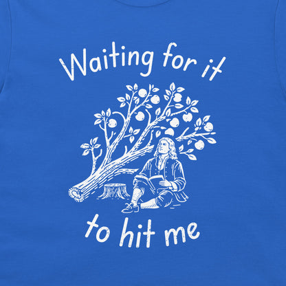 A close-up of a royal blue t-shirt featuring a white graphic print of a tree with fruit and a historical figure sitting beneath it, with the text 'Waiting for it to hit me' written in white script.