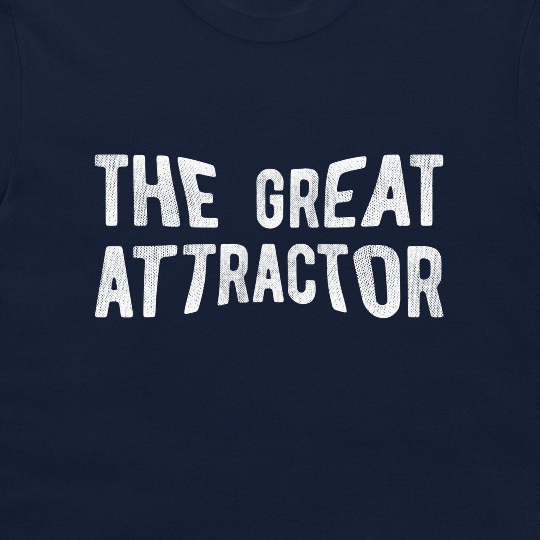 A close-up of a navy blue t-shirt featuring the phrase 'THE GREAT ATTRACTOR' printed in a distressed white typewriter-style font across the chest.