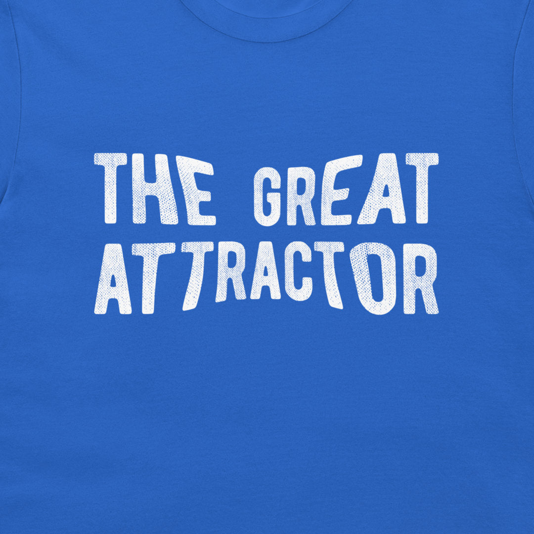 A close-up of a royal blue t-shirt featuring the phrase 'THE GREAT ATTRACTOR' printed in a bold, white, textured brush-style font across the chest.