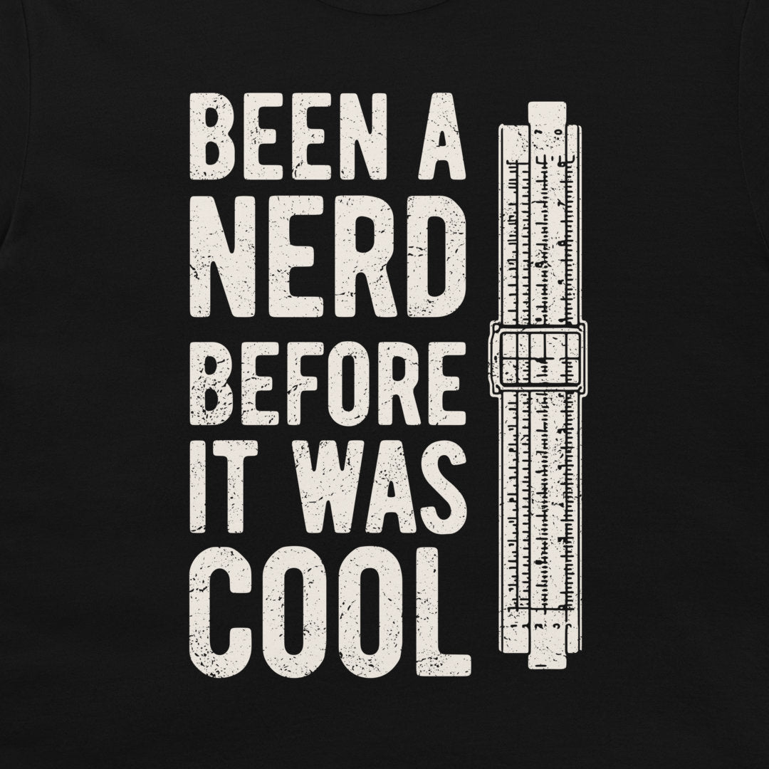 A black t-shirt featuring a distressed white graphic print that reads 'BEEN A NERD BEFORE IT WAS COOL' next to a vintage slide rule measuring device.