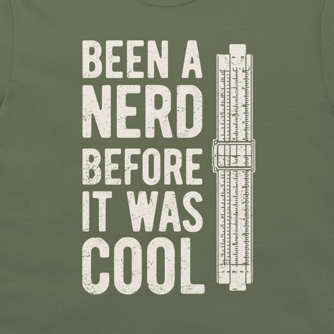 An olive green t-shirt featuring a distressed white graphic that reads 'BEEN A NERD BEFORE IT WAS COOL' next to a vintage slide rule measuring device.