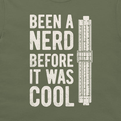 An olive green t-shirt featuring a distressed white graphic that reads 'BEEN A NERD BEFORE IT WAS COOL' next to a vintage slide rule measuring device.