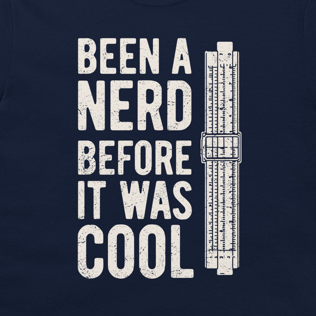 A close-up of a navy blue t-shirt, featuring a distressed white graphic print that reads 'BEEN A NERD BEFORE IT WAS COOL' next to a vintage slide rule measuring device.