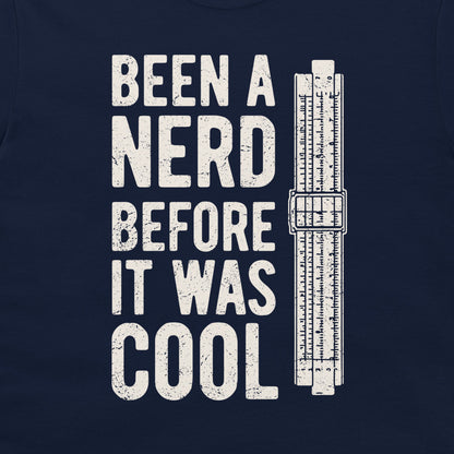 A close-up of a navy blue t-shirt, featuring a distressed white graphic print that reads 'BEEN A NERD BEFORE IT WAS COOL' next to a vintage slide rule measuring device.