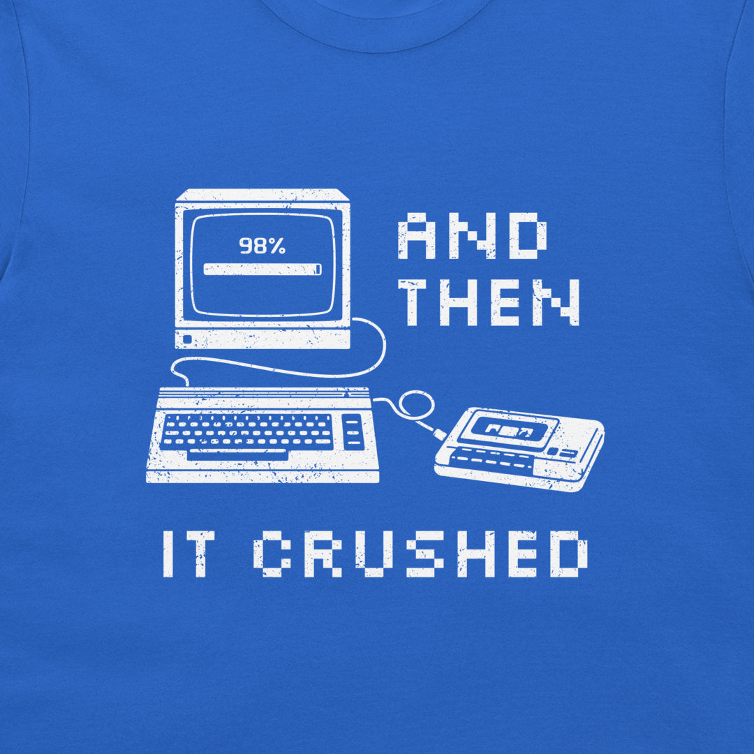 A close-up of a blue T-shirt featuring a white pixel-art graphic of a retro computer, keyboard, cassette tape, and the text 'AND THEN IT CRUSHED' in a distressed, retro computer-style font.