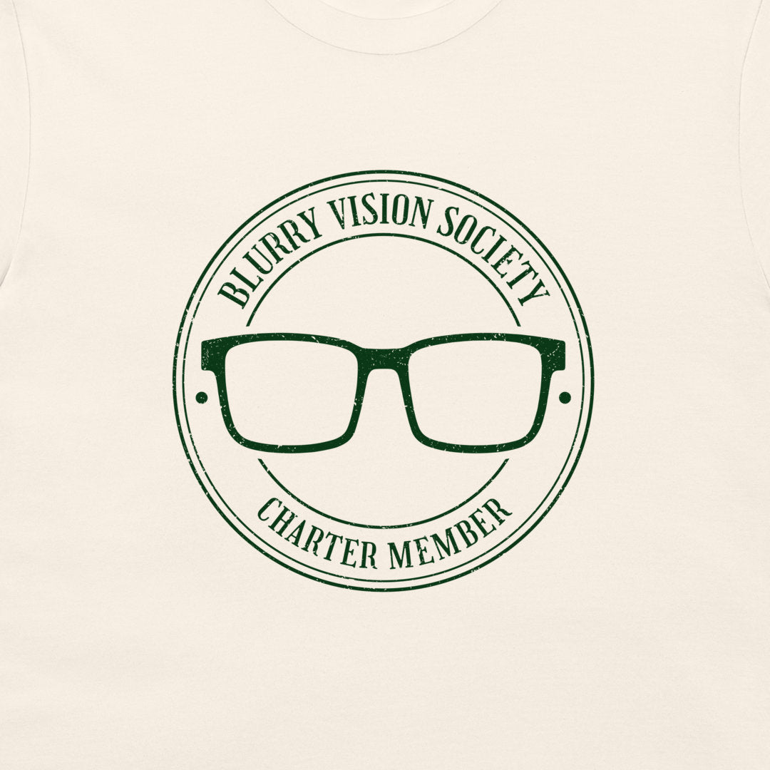 A close-up of a cream-colored t-shirt featuring a dark green circular graphic with the text 'BLURRY VISION SOCIETY CHARTER MEMBER' surrounding an illustration of eyeglasses.